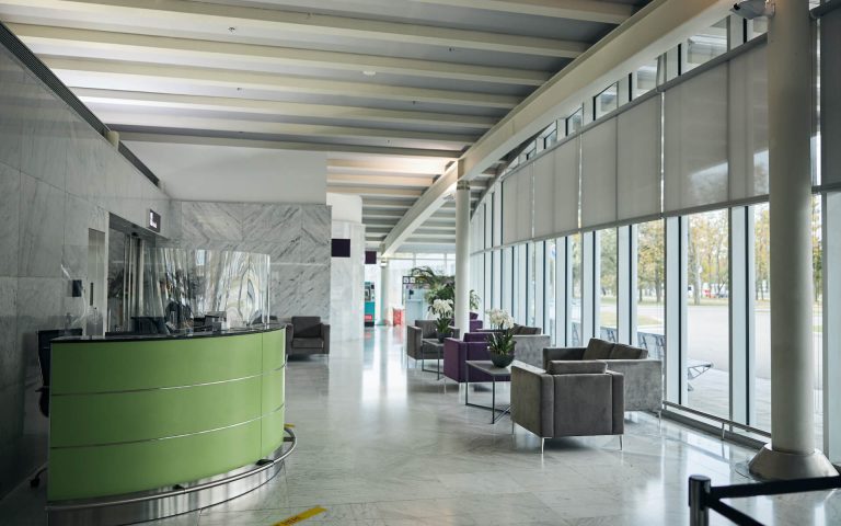 Moderne Lobby mit grünem Rezeptionstresen, grauen Stühlen, Marmorböden, großen Fenstern mit Jalousien und Zimmerpflanzen. Tageslicht durchflutet den großzügigen Innenraum – ein einladender Raum für ADHS-Coaching-Sitzungen in modernem Ambiente.