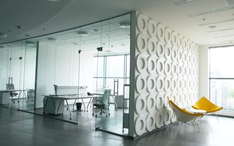 Moderne Büroräume mit verglasten Besprechungsräumen, weißen Schreibtischen und einer dekorativen Wand mit kreisförmigen Mustern – ideal für Coaching und Neurodivergenz. Zwei gelbe Stühle stehen neben großen Fenstern, die Tageslicht hereinlassen.